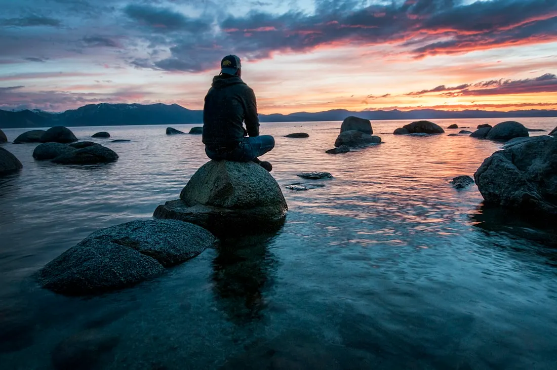 Person sitting quietly on a lakeside rock at sunrise